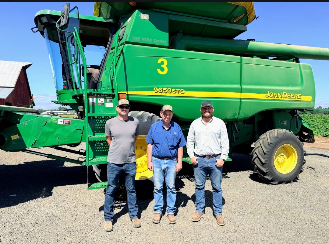 Three men stand in front of a large green John Deere 9650 STS combine marked with the number 3.